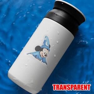 Puede incluir: Taza de viaje blanca con tapa negra, con una pegatina de Mickey Mouse con un atuendo de mago azul. La palabra "TRANSPARENT" est&aacute; impresa en rojo en la parte inferior de la taza.