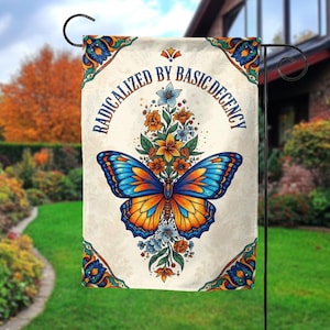 May include: A garden flag with a butterfly design. The butterfly has blue and orange wings, surrounded by flowers. The flag has the text "RADICALIZED BY BASIC DECENCY" in a semi-circular pattern. The flag is cream-colored with decorative corner designs.