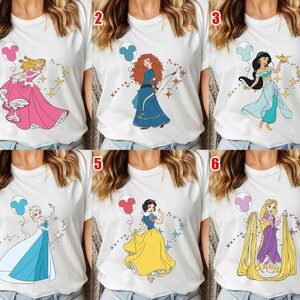 May include: White t-shirts featuring various cartoon princess designs. Each shirt showcases a different princess with a balloon and sparkling accents. The princesses include Aurora, Merida, Jasmine, Elsa, Snow White, and Rapunzel.
