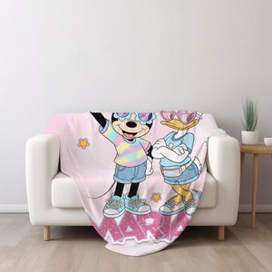 Op de afbeelding: Een roze sprei met een cartoon illustratie van Minnie Mouse en Daisy Duck met zonnebrillen en casual outfits. De sprei heeft de naam "MARIA" in roze letters en bloemaccenten.