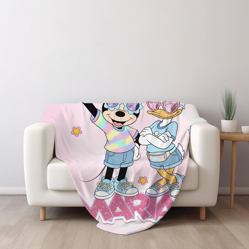 Op de afbeelding: Een roze sprei met een cartoon illustratie van Minnie Mouse en Daisy Duck met zonnebrillen en casual outfits. De sprei heeft de naam "MARIA" in roze letters en bloemaccenten.