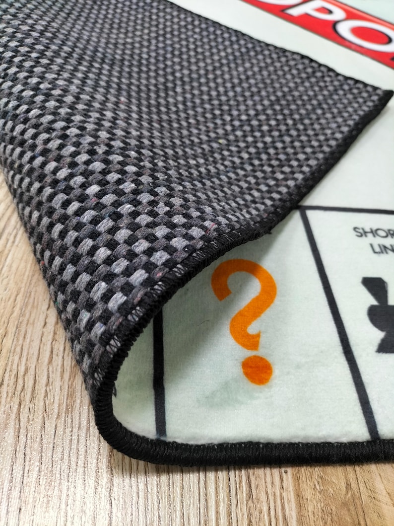 May include: A close-up of a Monopoly game board rug. The rug has a black and gray checkered pattern on the back. The front of the rug has a light green background with an orange question mark and black game board elements.