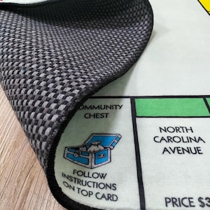 May include: A close-up of a Monopoly board game rug with a black and gray checkered backing. The rug features the Community Chest and North Carolina Avenue spaces. The text "Follow instructions on top card" is visible.
