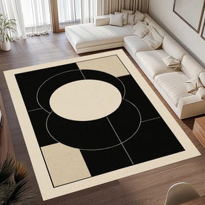 May include: A large, rectangular area rug with a modern geometric design. The rug features a black and cream color scheme with a central cream-colored oval shape, surrounded by black and cream geometric shapes and lines. The rug is placed on a hardwood floor.