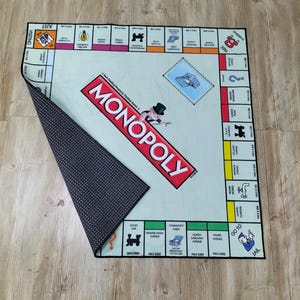 May include: A Monopoly board game mat with a light green playing surface and a black and white patterned underside. The Monopoly logo is prominently displayed in red. The board game's properties and spaces are visible.