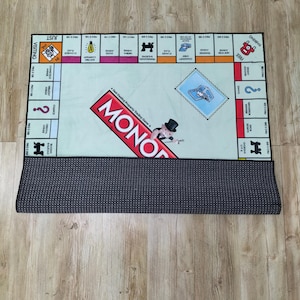 May include: A Monopoly board game rug with a light green center and a black border. The game board features property spaces, Chance and Community Chest cards, and the Monopoly logo. The rug is rolled up at the bottom, revealing a dark gray, dotted backing.