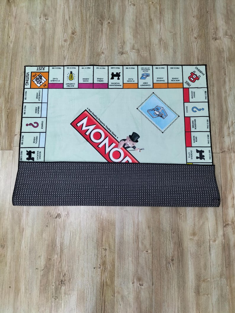 May include: A Monopoly board game rug with a light green center and a black border. The game board features property spaces, Chance and Community Chest cards, and the Monopoly logo. The rug is rolled up at the bottom, revealing a dark gray, dotted backing.