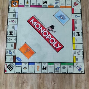 May include: A Monopoly board game rug with a light blue center and a red Monopoly logo. The game board features property spaces, Chance and Community Chest cards, and the Go to Jail space. The rug is designed for playing the classic board game.