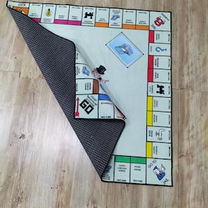 May include: A Monopoly board game rug with a light blue playing surface and colorful property spaces. The rug features the iconic Monopoly man and game board elements. The underside is a dark gray, dotted pattern.