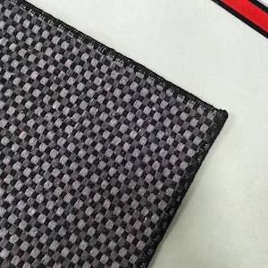 May include: Close-up of a section of a black and gray checkered rug with a black border. The rug is on a white surface, with a red and white graphic in the background.