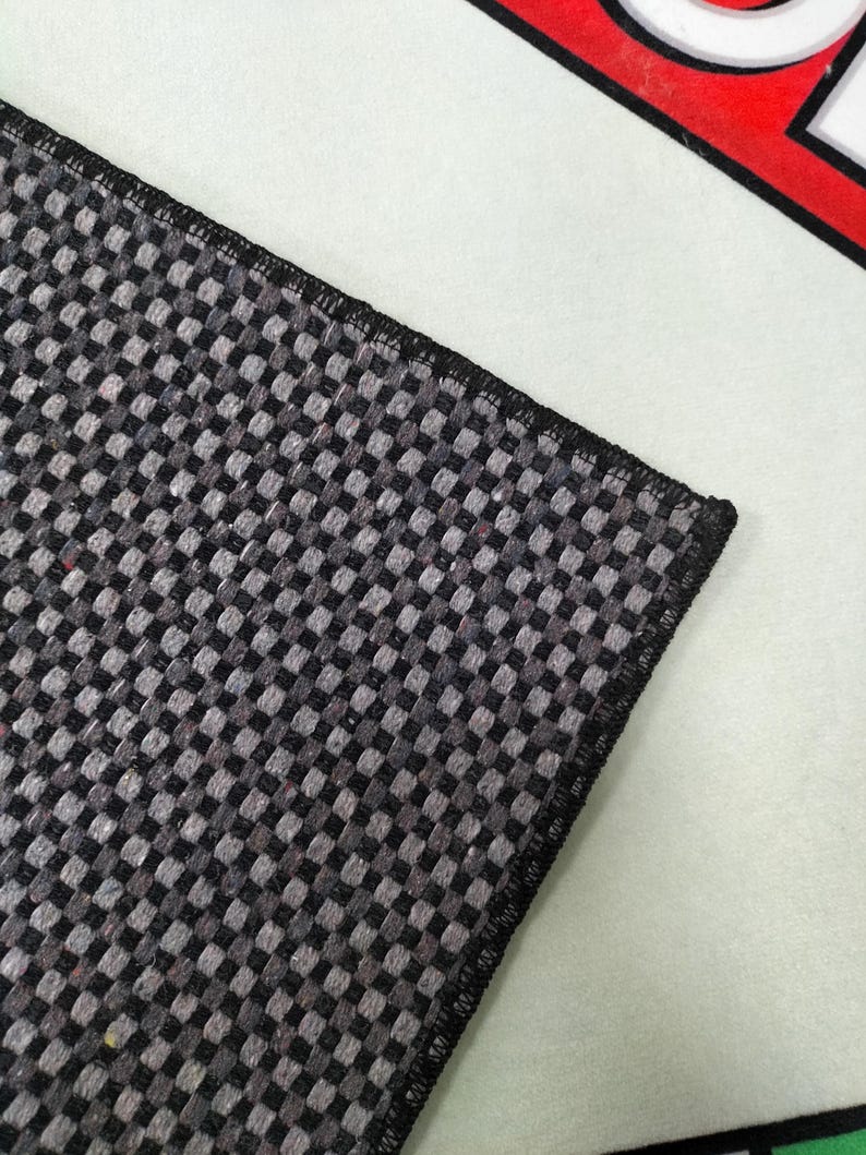 May include: Close-up of a section of a black and gray checkered rug with a black border. The rug is on a white surface, with a red and white graphic in the background.