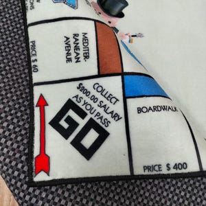 May include: A section of a Monopoly board game rug. The rug features the iconic Monopoly character, properties like Mediterranean Avenue, and the 'GO' space. The rug has a black border and is partially rolled up, revealing the game's design.