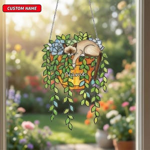 May include: A hanging decorative piece featuring a cat resting in a hanging planter with cascading green leaves and blue flowers. The planter has the word "Name" and a red banner with "CUSTOM NAME". The piece hangs from a silver chain.