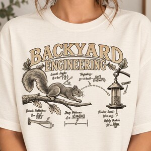 May include: An off-white t-shirt featuring a graphic print titled BACKYARD ENGINEERING. The design shows a squirrel on a tree branch, a bird feeder, and various physics equations including launch angle, trajectory, and feeder load calculations.