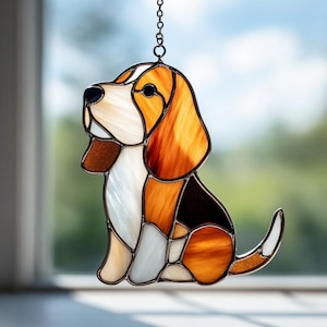 May include: A stained glass beagle dog suncatcher. The dog is depicted in a sitting position with a white, brown, and orange colour scheme. The suncatcher is hanging from a chain and is positioned in front of a window.