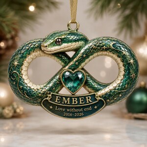 May include: A green and gold snake ornament shaped like an infinity symbol with a central heart-shaped green gemstone. A banner at the bottom features the text EMBER, Love without end, and 2016-2026. The snake has detailed scales and stars.
