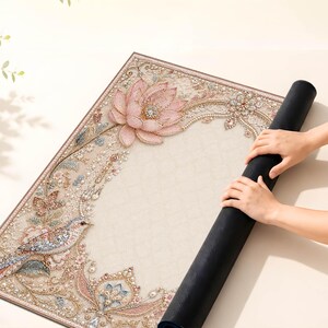 May include: A rectangular desk mat featuring a cream-colored surface with an intricate, sparkling design of a pink lotus flower, a small bird, and ornate floral filigree patterns in shades of pink, blue, and gold, with a solid black underside.