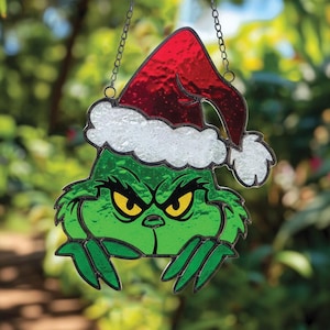 Op de afbeelding: Een gebrandschilderd glazen kerstornament met een groen personage met een boze uitdrukking, met een rode kerstmuts met witte rand. Het ornament hangt aan een zilveren ketting tegen een vage groene achtergrond.