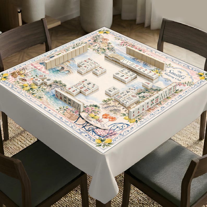 May include: A square mahjong table cover featuring a printed Mediterranean landscape with lemons, flowers, and a bistro table. The fabric displays the text "The Smith's Mahjong Club" and is arranged with several sets of white mahjong tiles.
