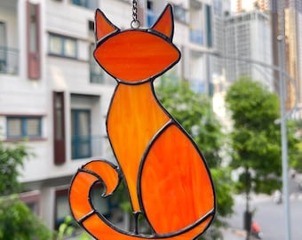 May include: A stained glass cat suncatcher in shades of orange and yellow, with black edging. The cat is depicted in a seated position with a curled tail. It hangs from a chain. The background shows buildings and trees.