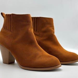 Könnte beinhalten: Ein Paar braune Wildlederstiefeletten mit Blockabsatz. Die Stiefeletten haben einen Pull-on-Stil und eine leicht spitz zulaufende Spitze.