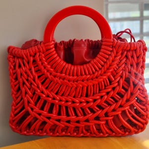 May include: A vibrant red handbag with a woven design. The bag features a circular handle and a drawstring closure. The woven pattern creates a unique texture, and the bag is made of a soft material.