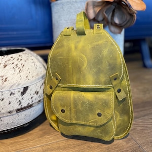 Pode incluir: Uma pequena mochila de couro amarela com um bolso frontal e fecho de aba. A mochila tem um visual envelhecido e é feita de couro de alta qualidade.