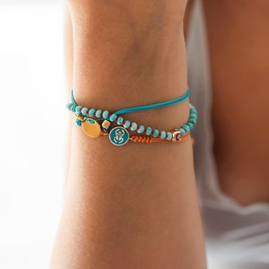 Può includere: Un braccialetto turchese e oro con un ciondolo che presenta un simbolo blu. Il braccialetto è fatto di perline e cordoncino.