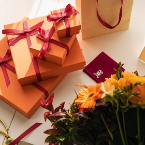 Puede incluir: Un conjunto de cajas de regalo naranjas atadas con una cinta color burdeos, una bolsa de terciopelo burdeos con un dije de mariposa plateado, un ramo de flores naranjas y amarillas y un par de tijeras doradas.
