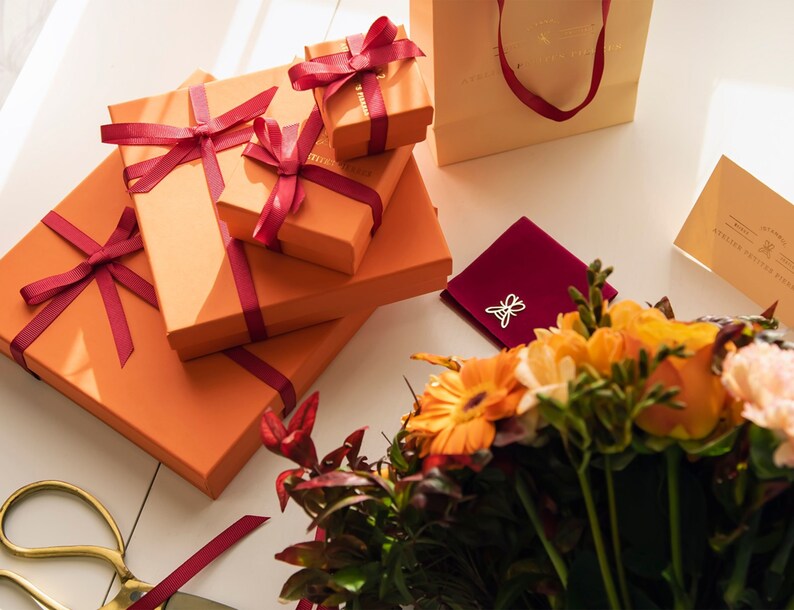 Puede incluir: Un conjunto de cajas de regalo naranjas atadas con una cinta color burdeos, una bolsa de terciopelo burdeos con un dije de mariposa plateado, un ramo de flores naranjas y amarillas y un par de tijeras doradas.