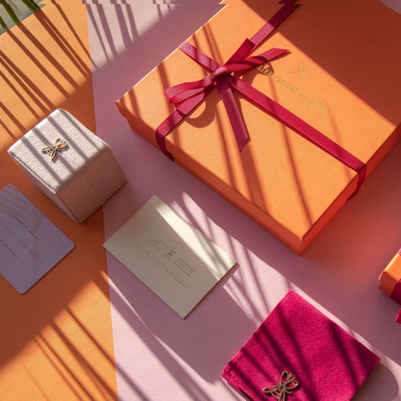 May include: A pink and orange background with a small beige box with a gold butterfly design, a white card with gold text that reads "Atelier des Petites Pierres", a larger orange box with a red ribbon and a gold butterfly design, and a small red velvet pouch with a gold butterfly design.