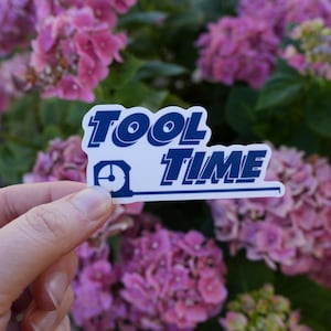 Puede incluir: Una pegatina blanca con texto azul que dice "TOOL TIME" con un icono de reloj. La pegatina está siendo sostenida por una mano frente a una flor rosa.