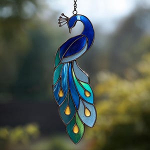 Könnte beinhalten: Ein Glasfenster-Pfau-Suncatcher mit einem dunklen Metallrahmen. Der Pfau hat Blau-, Türkis- und Grüntöne mit gelben Akzenten. Der Suncatcher hängt an einer Kette und zeigt die elegante Form des Vogels.