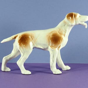 Puede incluir: Una figurita de porcelana blanca de un perro de caza marrón y blanco. El perro está de pie con la cabeza girada hacia un lado y la boca ligeramente abierta.