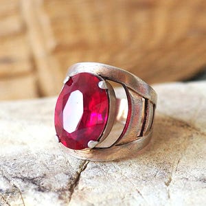 May include: A silver ring with a large, oval-cut, ruby-red gemstone. The ring has a wide band with a cut-out design on the sides. The gemstone is the focal point, catching the light and reflecting a vibrant color.