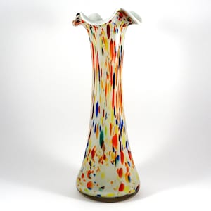Puede incluir: Un jarrón de vidrio blanco y alto con un borde ondulado. El jarrón está decorado con manchas de colores que parecen confeti en tonos de rojo, naranja, amarillo, azul y verde.