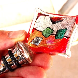 May include: A red glass bottle stopper with a silver and green geometric design. The stopper is attached to a silver metal base.