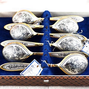 Op de afbeelding: Set van zes sierlijke zilveren lepels met goudkleurige accenten, gepresenteerd in een bruine doos met een blauw fluwelen interieur. Elke lepel heeft een gedetailleerd bloemenpatroon.