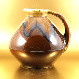Könnte beinhalten: Ein glänzender, brauner Keramikkrug mit gebogenem Henkel. Der Krug hat einen bauchigen Körper und einen leicht ausgestellten Rand. Die Glasur weist Brauntöne, Blau und Gold auf und erzeugt eine reflektierende Oberfläche. Der Krug ist ein dekorativer Gegenstand.