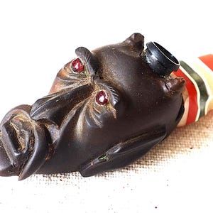 May include: A wooden pipe with a carved face and red gemstone eyes. The pipe is decorated with red, white and green stripes.