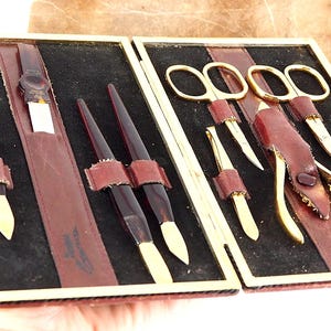 May include: An open vintage manicure set with a black velvet interior. The set includes various tools with gold-colored metal and dark brown handles, secured by leather straps. The case has a gold-colored frame.