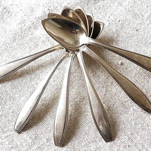 May include: A set of six silver teaspoons arranged in a fan shape on a white linen background.