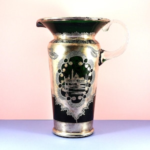 May include: An antique glass pitcher with a clear handle and a silver-colored base and rim. The pitcher has a dark green top and a silver-colored design around the body. The design includes a landscape scene and decorative patterns.