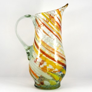 May include: A clear glass jug with a swirl design in shades of yellow, orange, and brown. The jug has a handle and a wide base.