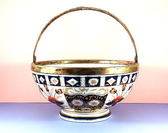 Könnte beinhalten: Ein dekorativer Keramikkorb mit goldenem Griff und Rand. Der Korb hat eine weiße Basis mit aufwendigen blauen, goldenen und roten Blumenmustern. Der Rand ist mit Gold verziert. Der Korb steht auf einem kleinen, runden Fuß.