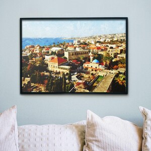 May include: Framed print of a coastal city with buildings featuring red tile roofs and a blue-domed structure. The artwork is set against a backdrop of a blue sea and sky, creating a scenic view. The print is displayed on a light blue wall.