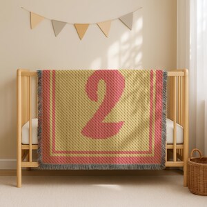 Könnte beinhalten: Eine gewebte Babydecke mit einer großen rosa Zahl 2 auf gelbem Hintergrund. Die Decke hat einen rosa Rand und Fransen. Die Decke ist über ein Holzbettchen drapiert.