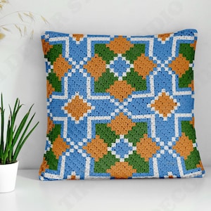 May include: A square crochet pillow cover featuring a geometric pattern in blue, green, orange, and white. The design consists of repeating diamond and cross shapes created with yarn, showcasing a textured, handmade stitch construction throughout.