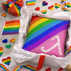 Könnte beinhalten: Ein gehäkeltes Kissen mit Regenbogenmuster in einer weißen Geschenkbox. Das Kissen zeigt ein diagonales Regenbogenmuster und ein weißes Symbol. Weitere Dekorationen im Regenbogen-Design sind eine Flagge, Herzen und ein Geschenk.