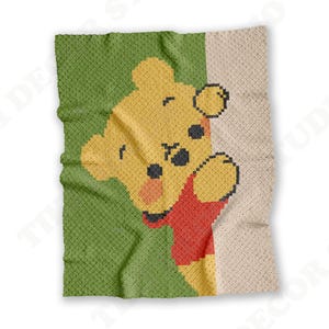 May include: A rectangular crochet blanket featuring a pixelated design of a yellow bear with a red shirt. The bear is positioned against a split background of solid green on the left and a textured off-white color on the right side of the blanket.